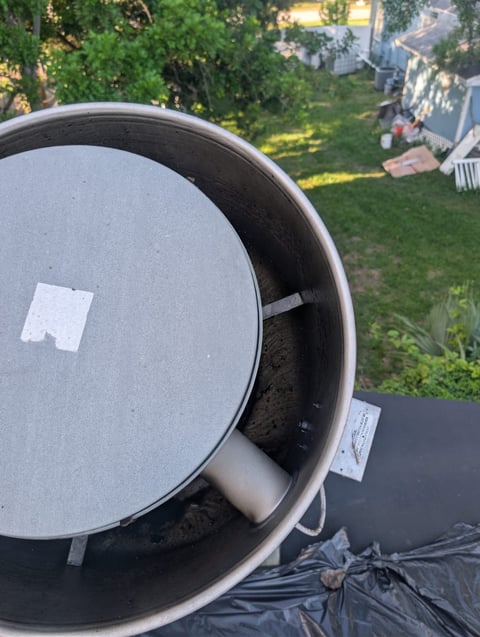 Overhead view of an open metal pot with gray lid sitting on black tarp in a backyard with house and green lawn visible